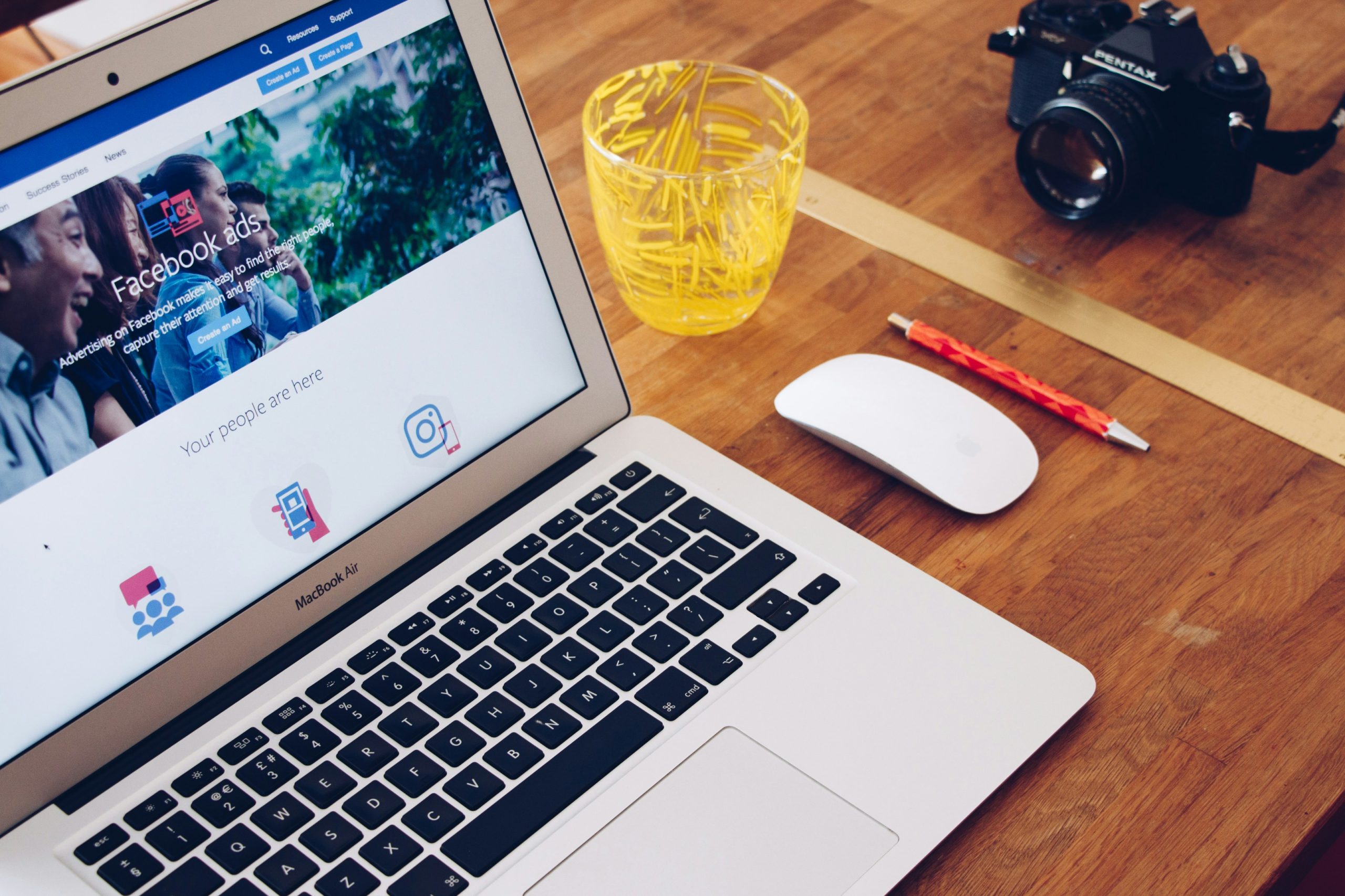 Ordinateur portable (macbook air) gris ouvert posé sur une table en bois à côté d'un verre jaune, d'une souris blanche connectée et d'un stylo rouge. Fenêtre Facebook Ads.