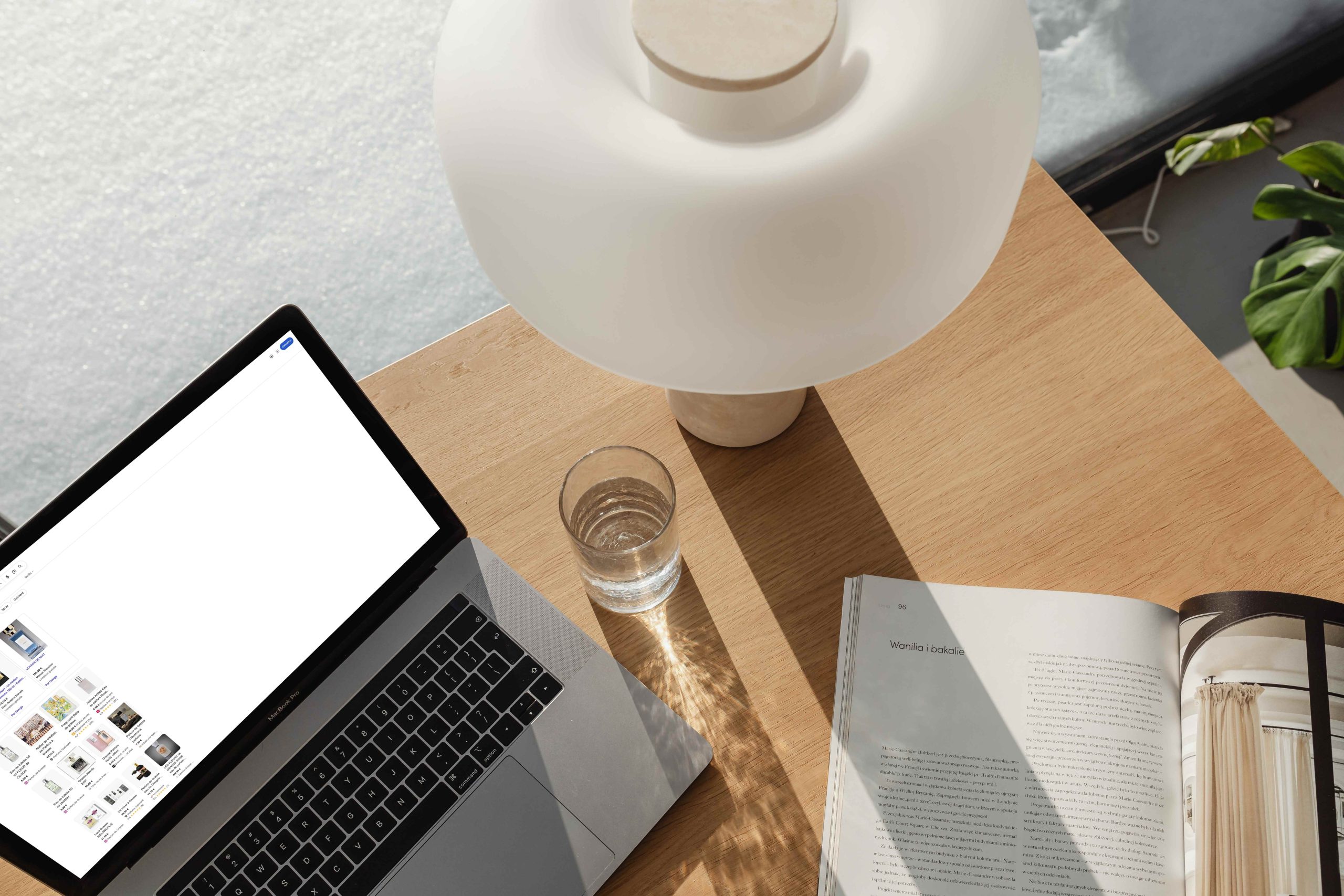 Bureau en bois avec un ordinateur portable ouvert affichant des vignettes de produits, un verre d’eau, une lampe blanche moderne et un magazine ouvert, éclairés par la lumière naturelle.