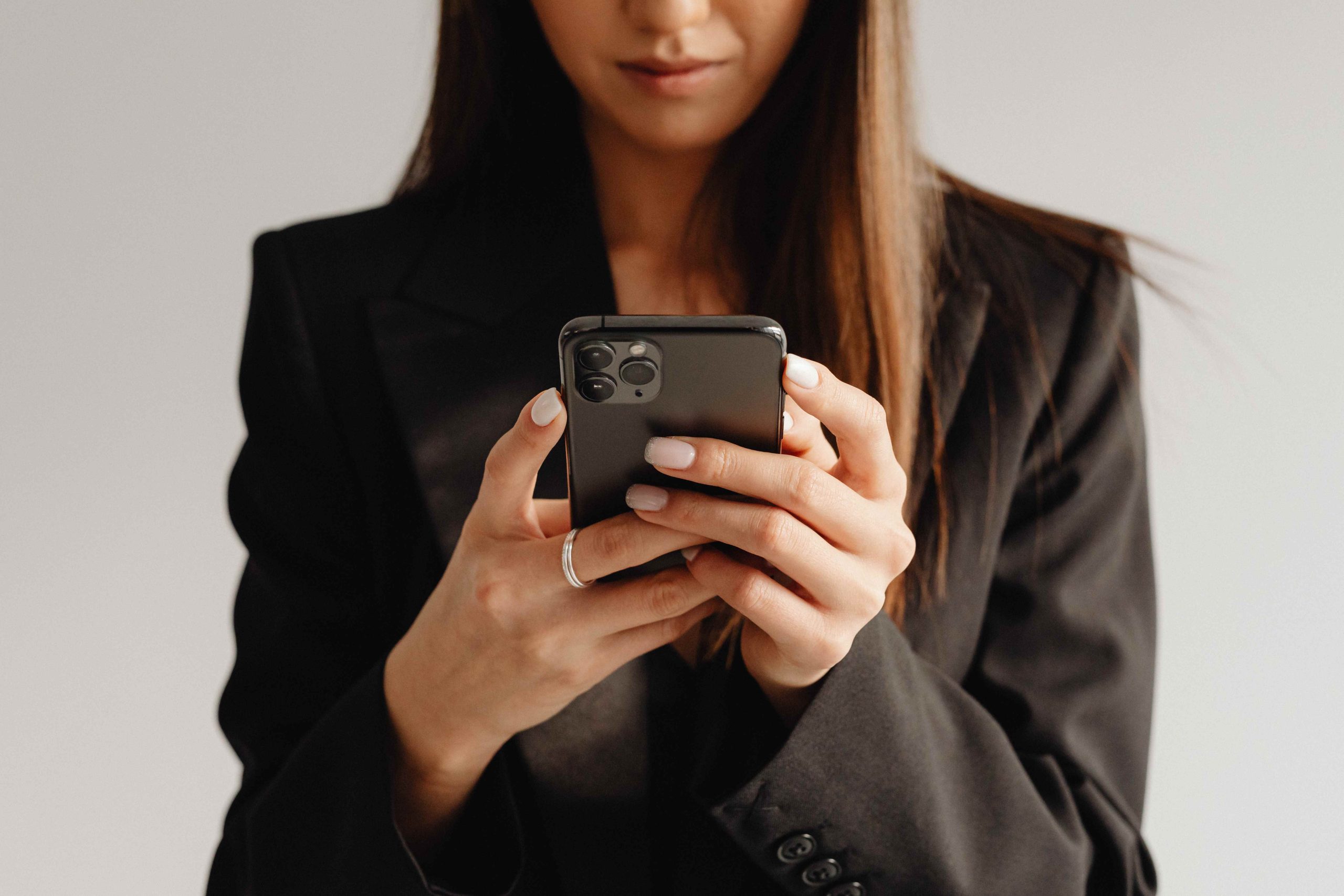 Une femme en blazer noir tient un smartphone à deux mains et regarde l’écran avec concentration.